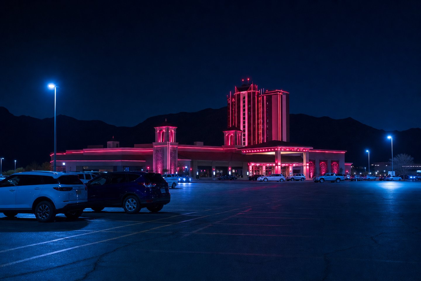 A casino resort seen at night from across its own parking lot, its facade edged in red neon, with low mountains as a dark silhouette on the horizon behind it.